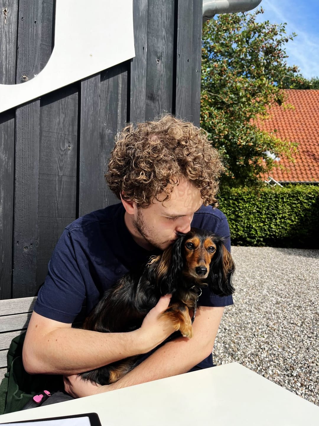 Nicolai, curly-haired in a navy T-shirt, kisses the head of Frida, a long-haired black and tan dachshund, held close on a sunny bench.